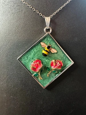 Halsband med bi och rosor - Unikt halsband med fyrkantigt hänge i silverfärg. Hänget har en grön bakgrund med detaljerade röda rosor och ett bi i gult och svart. Kedjan är tunn och elegant. Perfekt för dig som gillar naturinspirerade smycken och vill sticka ut med något färgglatt och lekfullt.