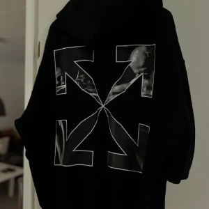 Svart hoodie från Off-White - Svart hoodie från Off-White med ikoniskt piltryck på ryggen och vit grafisk detalj. Klassisk huva och mjukt material, perfekt för en streetwear-look. Snygg och stilren design som sticker ut. Galet bra pris pga flytt. Passa på och göra en fantastisk affär. 