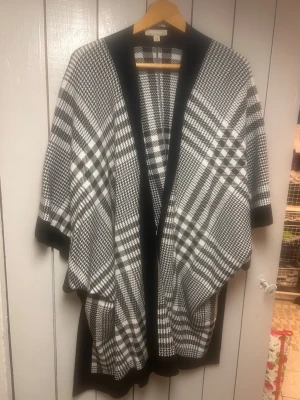 Svartvit rutig poncho från Barbour - Snygg svartvit poncho från Vero Moda med klassiskt rutigt mönster. Öppen framtill och bred passform, perfekt att slänga över en tröja. Tjockt, mjukt material och svarta kanter för en stilren look.