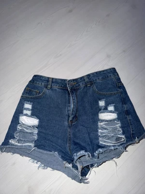 Blå denimshorts med slitningar - Snygga blå denimshorts med hög midja och råa, fransiga kanter. Shortsen har slitna detaljer framtill och klassisk femficksdesign. Perfekta för en avslappnad och trendig look. Storlek L