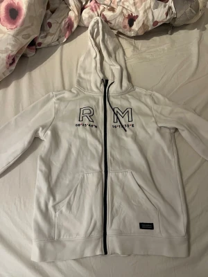 Vit hoodie med dragkedja - Vit hoodie med svart dragkedja och huva. På bröstet finns tryck med bokstäverna 'RM' och koordinater under. Två fickor framtill och en liten svart logotyp-patch nertill. Perfekt för en chill och avslappnad stil.