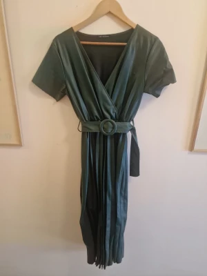 Grön omlottklänning med bälte - Snygg lång grön klänning i läderimmitation från New Collection med korta ärmar och matchande bälte i midjan. Klänningen har v-ringning och ett elegant fall. Perfekt för dig som gillar stilrena och trendiga plagg.