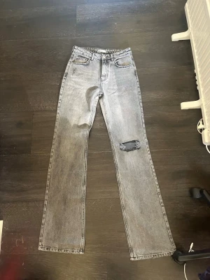 Grå raka jeans med slitning - Säljer ett par grå raka jeans med en snygg slitning på ena knät. Jeansen har klassisk femficksdesign och hög midja. Perfekta för dig som gillar en avslappnad och trendig look. Materialet är denim i bomull. Lite långa i benen.  Kan diskutera pris