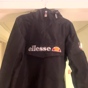 Svart vindjacka från Ellesse - Snygg svart vindjacka från Ellesse med stor logga på bröstet och patch på ärmen. Jackan har huva med dragsko och en praktisk magficka med lock. Perfekt för dig som vill ha en sportig och stilren look. Materialet är tunt och vindtätt.