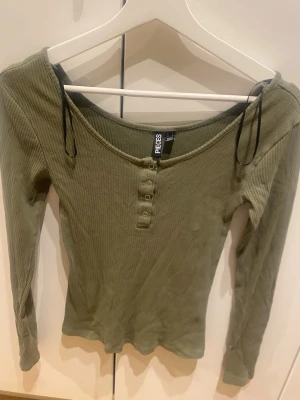 Olivgrön ribbad topp från Pieces - Snygg olivgrön ribbad topp från Pieces med lång ärm och knappar framtill. Toppen har en djupare rundad halsringning och är tillverkad i mjukt material som sitter skönt på kroppen. Perfekt att styla med jeans eller kjol.