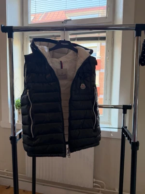 Svart dunväst från Moncler med huva - Snygg svart dunväst från Moncler med vit insida och huva. Västen har två dragkedjefickor framtill, quiltad design och klassisk Moncler-logga på bröstet. Perfekt för lager-på-lager och streetstyle.
