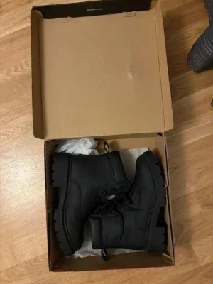 Svarta snörade stövletter - Säljer ett par svarta stövletter med grov sula och snörning framtill. Skorna har en klassisk kängdesign med vadderad kant och robust yttersula. Perfekta för dig som gillar en tuffare stil. Materialet ser ut att vara syntet eller läderimitation.