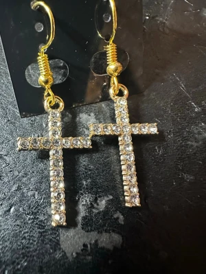 Guldiga korsörhängen med strass - Snygga örhängen i form av kors med gnistrande klara strasstenar. Örhängena är guldfärgade och har krokfäste. Perfekta för dig som vill ha ett statement-smycke med lite extra bling.