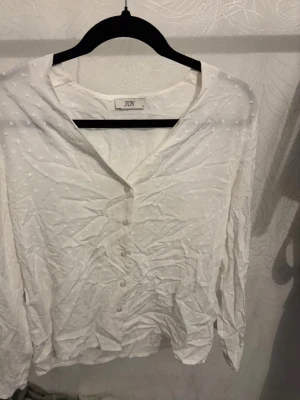 Vit blus med knappar från JDY - Snygg vit blus från JDY med diskreta prickar i tyget och knäppning framtill. Blusen har v-ringning och långa ärmar. Perfekt att styla med jeans eller kjol för en fräsch look.