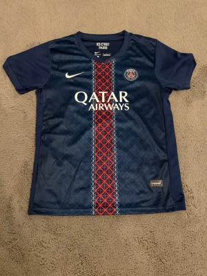 PSG matchtröja O. Dembélé #10 Nike - Mörkblå PSG matchtröja från Nike med röd och vit detalj i mitten, Qatar Airways-tryck och klubbmärke på bröstet. På ryggen står O. Dembélé och nummer 10. Perfekt för fotbollsfans som vill visa sitt stöd för Paris Saint-Germain.
