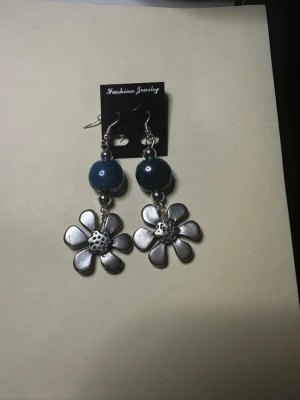 Blommiga örhängen med blå pärlor - Snygga örhängen med stora blå pärlor och hängande blommor i silverfärgad metall. Perfekt accessoar för att ge din stil en unik touch. Krokar i metall och cool design med tydlig blomform.