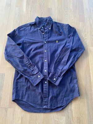 Marinblå skjorta från Ralph Lauren - Klassisk marinblå skjorta från Ralph Lauren med button-down krage och broderad gul logga på bröstet. Skjortan har långa ärmar, vita knappar och är tillverkad i bomull. Perfekt för en stilren och avslappnad look.