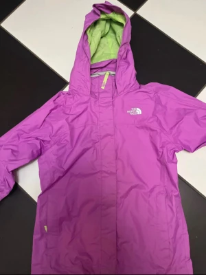 Lila regnjacka från The North Face - Cool lila regnjacka från The North Face med huva och ljusgrönt meshfoder. Jackan har dragkedja och logga på bröstet. Perfekt för att hålla sig torr när det regnar. Materialet är syntet och jackan har en sportig look.