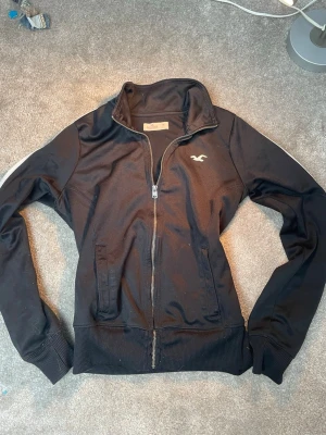 Svart hoodie från Hollister - Svart hoodie från Hollister med dragkedja. Klassisk logotyp på bröstet och två fickor. Materialet är mjukt och bekvämt, perfekt för en avslappnad stil. Kolla in mina andra annonser!🥰