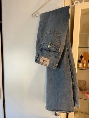 True Religion Becca Bootcut Jeans - Säljer ett par ljusblå bootcut jeans från True Religion, modell Becca. Jeansen har klassiska kontrastsömmar, snygga fickdetaljer med lock och silverfärgade knappar. Tillverkade i denim med en autentisk tvättad look.