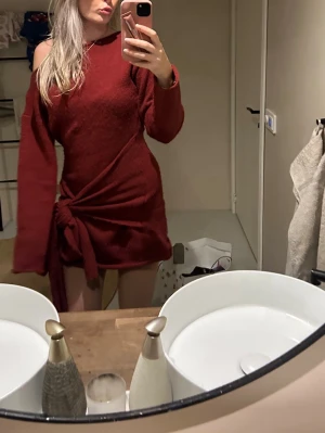 Vinröd omlottklänning med knyt - Vinröd kort omlottklänning med lång ärm och knyt i midjan. Klänningen har en offshoulder-design och är tillverkad i ett mjukt, stickat material som ger en mysig känsla. Perfekt för dig som vill ha en trendig och bekväm look.