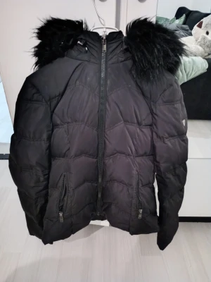 Svart pufferjacka med fuskpäls - Säljer en svart pufferjacka från saki med dragkedja framtill, två fickor med dragkedja och huva med svart fuskpäls. Jackan har ett diskret logomärke på ärmen och är perfekt för kalla vinterdagar. Materialet är syntet och jackan är riktigt fluffig och varm.
