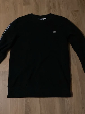 Svart Lacoste tröja - Svart sweatshirt från Lacoste med klassisk krokodillogga på bröstet och texten 'Lacoste' tryckt längs båda ärmarna. Tröjan har rund halsringning och är tillverkad i mjukt material, perfekt för en avslappnad streetwear-look.