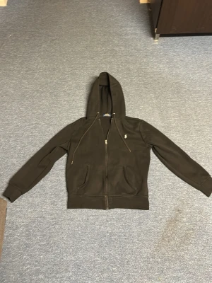 Svart hoodie från Polo Ralph Lauren - Säljer en svart hoodie från Polo Ralph Lauren med dragkedja framtill och två fickor. Klassisk broderad logga på bröstet och justerbar huva med snören. Tillverkad i mjukt bomullsmaterial som är skönt att ha på sig.