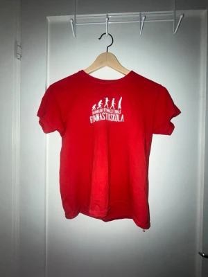 Röd t-shirt Gymnastikskola - Röd t-shirt med tryck på bröstet där det står 'Nynäshamns Gymnastikskola' och siluetter av gymnaster. Klassisk rund hals och korta ärmar. Perfekt för dig som gillar gymnastik och vill ha en enkel och sportig look.