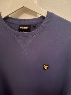 Blå sweatshirt från Lyle & Scott - Snygg blå sweatshirt från Lyle & Scott med rund halsringning och det klassiska gula fågel-logotypen broderad på bröstet. Tröjan har ribbade muddar vid ärmslut och nederkant samt en relaxed passform. Perfekt för en avslappnad stil.