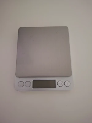 Digital gramvåg i silver - Säljer en digital köksvåg i silverfärgad metall och plast. Vågen har en platt, fyrkantig design med fyra knappar och en digital display framtill. Perfekt för att väga ingredienser noggrant i köket.