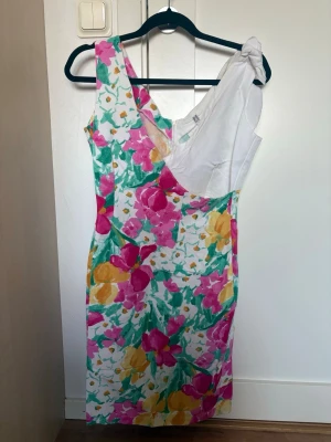 Färgglad blommig klänning  - Snygg kortklänning med färgglatt blommönster. Klänningen har en unik one shoulder-design med en rosett på axeln och är perfekt för sommarens tillfällen.