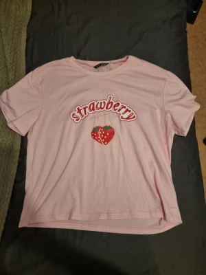 Rosa t-shirt med jordgubbstryck - Söt rosa t-shirt med trycket 'Strawberry' och en jordgubbe på bröstet. T-shirten har rund hals och korta ärmar. Perfekt för dig som gillar gulliga och lekfulla plagg. Någorlunda oversized 