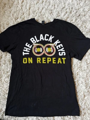 The Black Keys svart t-shirt - Svart t-shirt med tryck från The Black Keys. Framsidan har texten 'The Black Keys On Repeat' med färgglada vinylskivor, baksidan listar turnédatum och städer. Klassisk passform och rund hals. Perfekt för fans av bandet.