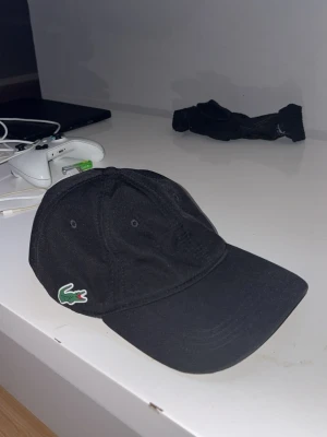 Svart keps från Lacoste - Snygg svart keps från Lacoste med klassisk böjd skärm och broderad grön krokodil-logga på sidan. Kepsen har sex paneler och justerbar rem baktill. Perfekt för en stilren och sportig look.