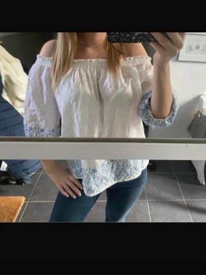 Vit offshoulder blus med blå brodyr - Säljer en söt vit offshoulder blus med blå broderade detaljer längs ärmslut och nederkant. Blusen har trekvartsärm och resår upptill för en bekväm passform. Perfekt till jeans eller kjol för en fräsch look.