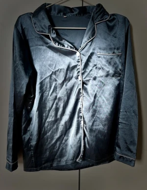 Blå satinpyjamas  - Snygg mörkblå pyjamas i satin med långärmad skjorta och matchande byxor. Skjortan har vita knappar framtill, kontrasterande vita passpoaler och en bröstficka. Materialet är glansigt och mjukt 💙 (180 kr för både byxa och tröja!)
