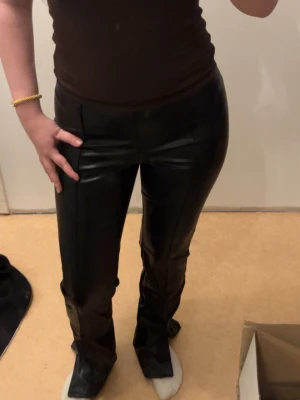 Svarta fuskskinnbyxor med pressveck - Stilrena svarta byxor i fuskskinn med pressveck framtill och utsvängda ben. Byxorna har hög midja och stängs med dragkedja i sidan. Perfekta för dig som vill ha en trendig och edgy look.