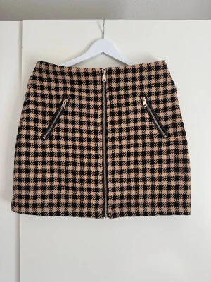 AMISU svart och beige mini kjol, storlek EU40 - Kort kjol med dragkedja framtill och två dekorativa dragkedjor på sidorna. Kjolen har ett rutigt mönster i svart och beige. Length: 46 cm   Buy shipping label or pick it up from Farsta Centrum in Stockholm.