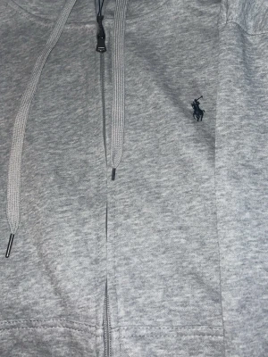 Grå hoodie från Polo Ralph Lauren - Snygg grå hoodie från Polo Ralph Lauren med dragkedja framtill och justerbar huva. Klassisk logga broderad på bröstet i mörkblått. Tillverkad i mjukt bomullsmaterial som känns skönt mot huden. Perfekt för en avslappnad och stilren look.
