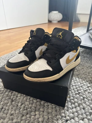 Air Jordan 1 Mid svart/vit/guld - Säljer ett par Air Jordan 1 Mid sneakers i svart, vitt och guld. Klassisk siluett med svart snörning, vit tåbox och guldfärgade detaljer som swoosh och logga. Ovandelen är i skinn och skon har en bekväm, vadderad krage. Box medföljer.