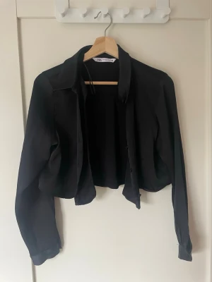 Svart croppad skjorta från Zara - Säljer en svart croppad skjorta från Zara med klassisk krage och långa ärmar. Skjortan har knappar framtill och är tillverkad i ett mjukt, tunt material som ger en snygg och avslappnad look.
