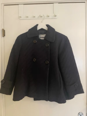 Mörkblå kappa från Intrend - Stilren mörkblå kappa från Intrend i ullblandning med dubbelknäppning framtill och dekorativa knappar på ärmslut och rygg. Kappan har en klassisk krage och en något utsvängd, kortare modell som ger en trendig siluett. Lite smått randig. 