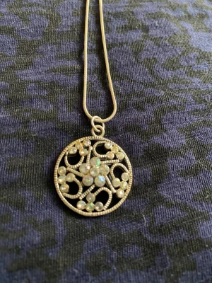 Silverfärgat halsband med pärlor - Snyggt halsband med en rund, silverfärgad hängare dekorerad med små pärlor och glittrande stenar i ett blommigt mönster. Kedjan är tunn och elegant, perfekt för att ge en extra touch till din outfit.