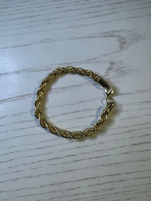 Guldfärgat tvinnat armband - Snyggt armband i guldfärgad metall med tvinnad design. Armbandet har ett klassiskt lås och passar perfekt för dig som gillar stilrena accessoarer med lite extra glans. Perfekt att bära ensam eller att matcha med andra smycken.