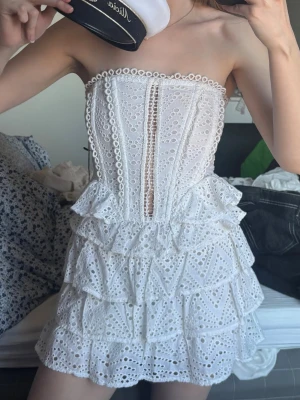Vit volangklänning - studentklänning  - Superfin vit kortklänning med volanger och broderade spetsdetaljer. Axelbandslös modell med dekorativa hålmönster. Som visas på bild håller tyget på att gå lite sönder fram