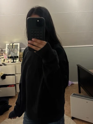 Svart hoodie i oversized modell - En svart hoodie med oversized passform och långa ärmar. Klassisk design utan synliga loggor eller mönster, perfekt för en avslappnad och cool look. Materialet ser ut att vara mjukt och bekvämt, idealiskt för chill dagar.