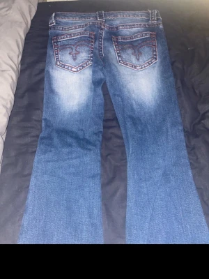 Blå bootcut jeans från Gina tricot - Säljer ett par blå bootcut jeans från True Religion med snygga slitningar och kontrastsömmar. Jeansen har broderade bakfickor och klassisk femficksdesign. Perfekta för dig som gillar en avslappnad men trendig stil.