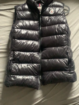Svart dunväst från Moncler - Snygg svart dunväst från Moncler med glansig finish och klassisk Moncler-logga på bröstet. Skulle vilja byta mot ett par skor.Kom med förslag på vilka skor och visa bild så diskuterar vi.Annars säljer jag för 1000kr går att pruta.