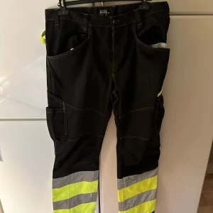 Svarta arbetsbyxor från Bluewear - Säljer ett par robusta svarta arbetsbyxor från Bluewear med reflexdetaljer i gult och grått. Byxorna har flera praktiska fickor och förstärkningar för extra hållbarhet. Perfekta för arbete där synlighet är viktigt.