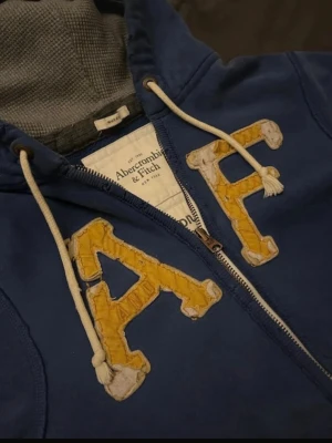 Blå hoodie från Abercrombie & Fitch - Blå zip-hoodie med stora gula bokstäver 'A F' på bröstet. Mjuk insida i grått tyg, vita dragsnören och kängurufickor framtill. Perfekt för en avslappnad och cool stil. Har cool disressting och är från 2000 talet.
