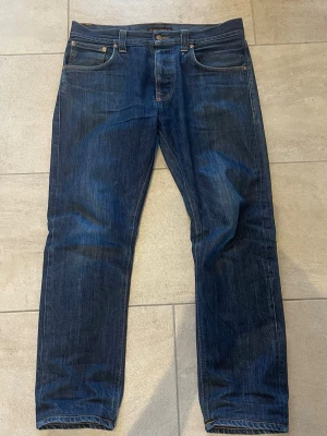 Mörkblå raw denim jeans från Nudie Jeans - Snygga mörkblå jeans från Nudie Jeans med klassisk femficksdesign och orangea sömmar på bakfickorna. Raka ben och normal midja. Tillverkade i slitstark denim av bomull. Perfekta för en avslappnad och stilren look. Jeans är discountied i modellen big Bengt och år 32/32 i storlek