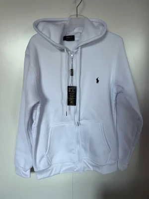 Vit zip hoodie från Polo Ralph Lauren i strl S - Snygg vit hoodie med dragkedja från Polo Ralph Lauren. Klassisk design med huva, två stora fickor framtill och den ikoniska lilla broderade loggan på bröstet. Tillverkad i mjukt fleece material som känns skönt mot huden. Perfekt för en clean och stilren look.Den är tyvärr inte äkta men fortfarande stilig ,varm och snygg.   Helt ny .Djur och rökfritt hem 