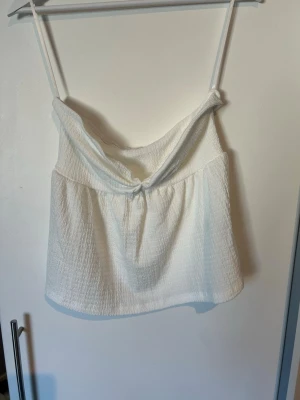 Vit tubtopp med extra detaljer - Säljer en vit tubtopp med snygg knytning framtill. Toppen har en lätt strukturerad yta och avslappnad passform, perfekt för varma dagar. Enkel och stilren design som är lätt att matcha.
