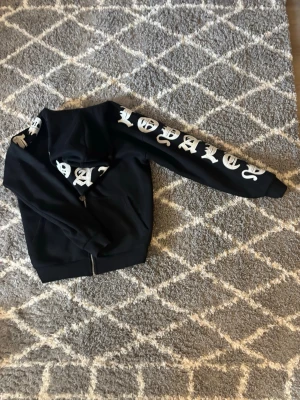 Svart hoodie från Black Squad S - Svart hoodie från Black Squad med dragkedja och stor vit text 'LOYALTY' på ena ärmen. Klassisk huva och fickor framtill. Perfekt för dig som gillar streetwear och vill sticka ut med coola detaljer.